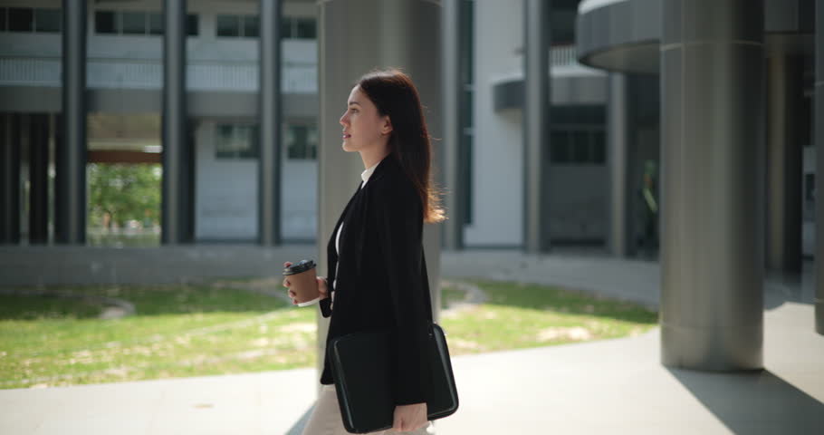 Side view shot, Footage of Young smiling elegant Asian busy business woman in a suit holding a cup of coffee and walking in front of a modern business building. Business and people concepts.