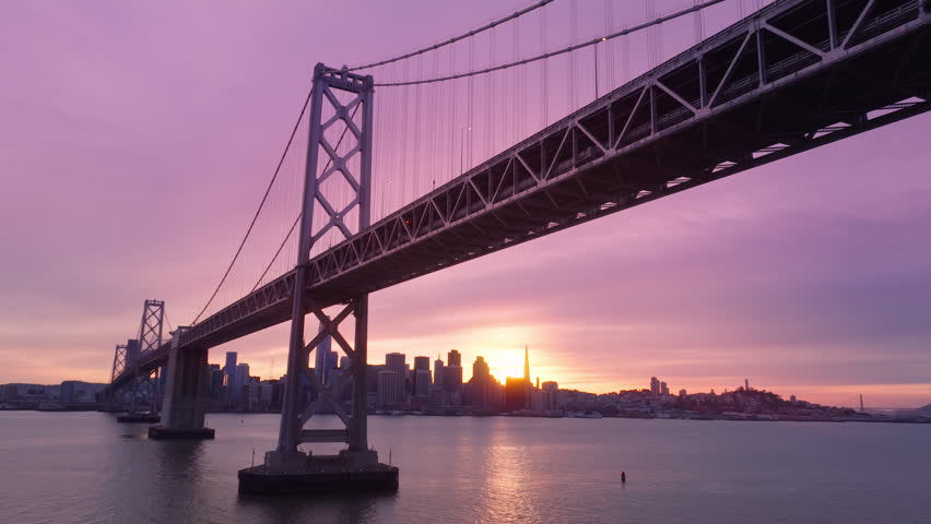 Drone flying above water surface under Bay Bridge construction with scenic cityscape silhouette on background. Beautiful aerial skyscrapers in downtown San Francisco in cinematic pink cloudy sky 4K