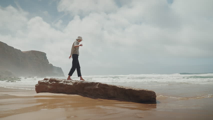 Young tourist woman with hat enjoying and having fun at the sandy beach by the ocean. Foamy sea waves are rolling towards the beach. Celebrating summer vacation. 