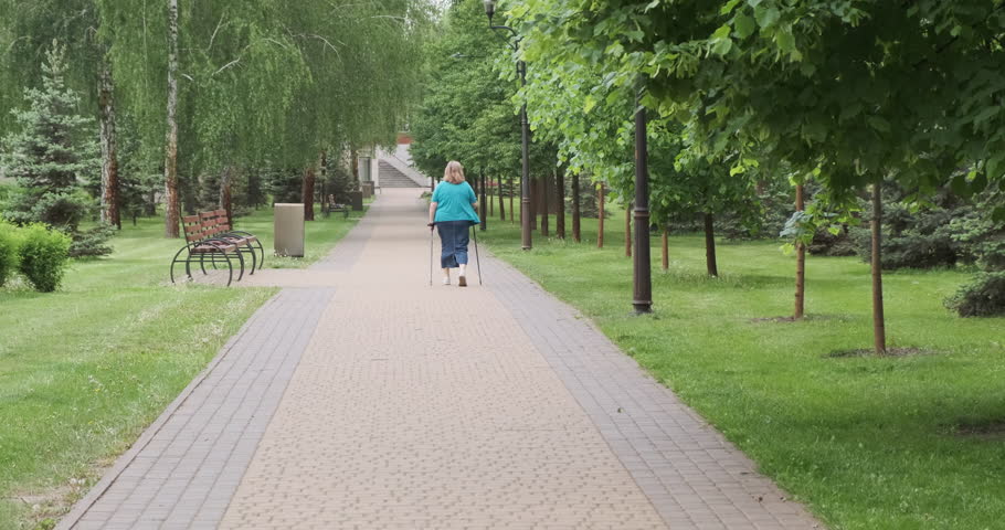 Unrecognizable woman walks with walking sticks in spring park. Back view following. Concept of physical exercise for health for the elderly, rehabilitation.