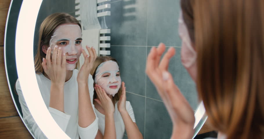 Portrait of two beautiful girls with facial face masks cosmetic looking at the mirror while standing in the bathroom at home. Skin care and beauty concept