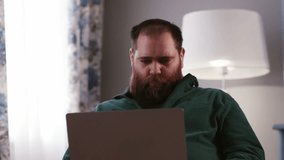 A frowning bearded man sits in front of a laptop. A male freelancer in a green sweatshirt works remotely at a laptop while sitting at home.