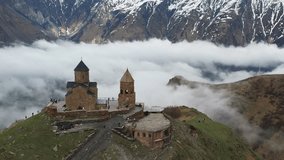 Aerial drone view of Gergeti Trinity Church with heavy fog clouds flowing over the mountain. Beautiful mountain view of the snow-capped peaks near Stepantsminda, Georgia.