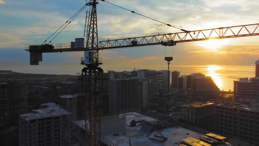 New modern apartment buildings, high-rise building with construction crane against backdrop of sunset, sun on ocean or sea. Drone view of crane construction site over city at sunset. Contractor. Royalty-Free Stock Footage #1104202689