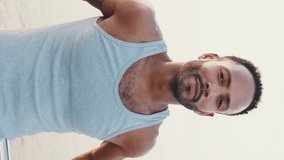 VERTICAL VIDEO: Young bearded male fit athlete standing on the embankment against the sea background, looking at the camera and smiling. Camera moving forwards