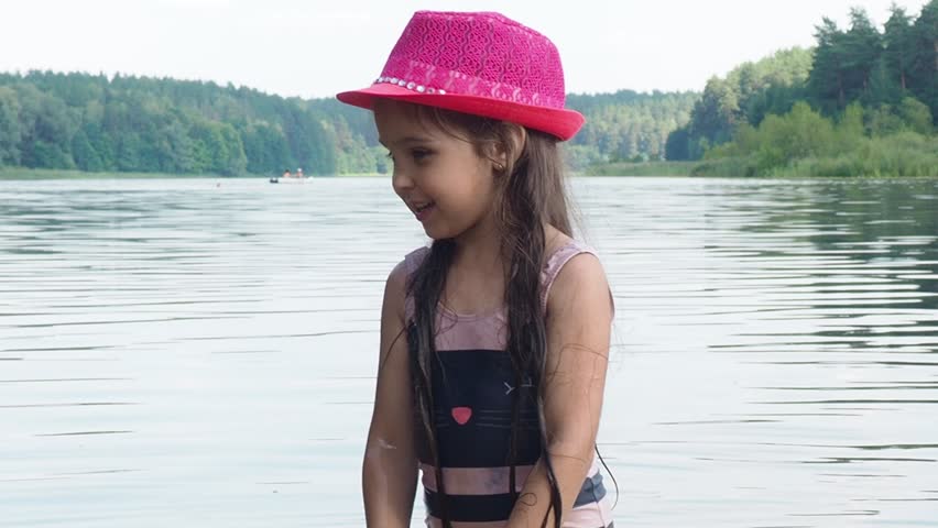 little girl smiling playing in the river or lake or sea. A girl with in a pink hat and long dark hair raises her hands up in the water and splashes water drops. laughing, smiling, having fun. summer 