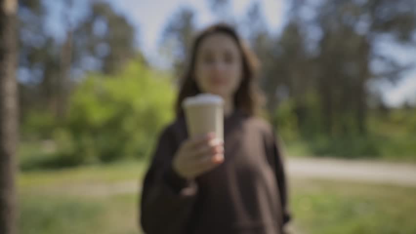 A girl shows a paper glass with a drink to the camera in the park. A woman hand holds a brown paper coffee cup with a white lid on a natural background. Woman hand holding brown paper coffee cup. Royalty-Free Stock Footage #1104209541
