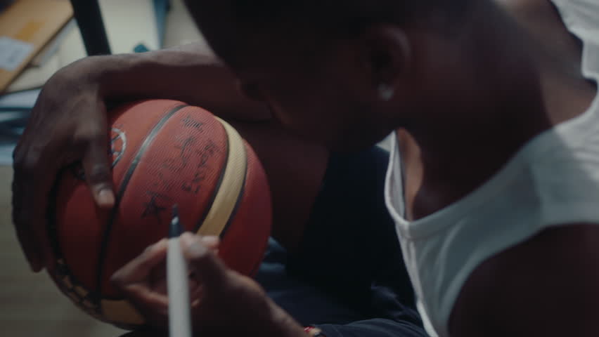 Young black basketball player sitting on couch at home, writing a sign on a ball with marker pen, making memorabilia. High angle view, medium close-up shot Royalty-Free Stock Footage #1104210563