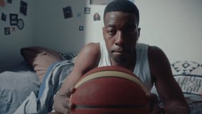 Young black male athlete holding basketball, sitting on couch at home, posing for camera with confidence. Medium shot, video portrait