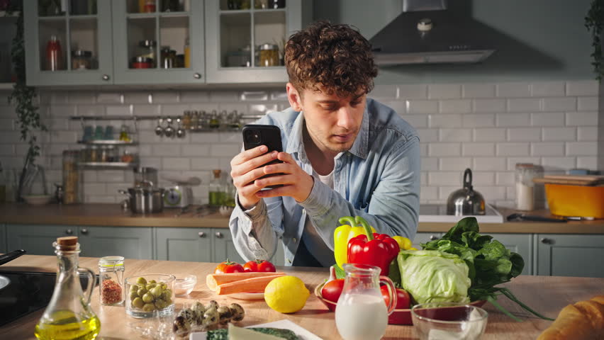 Young man standing by kitchen table with fresh healthy vegetables, using mobile phone, searching recipe for preparing yummy meal. Inspired chef amateur expressing happy emotions while cooking at home Royalty-Free Stock Footage #1104213147