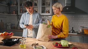 Beautiful young couple in love, unpacking a bag with fresh vegetables, checking the shopping list on smartphone, smiling, expressing positive emotions, standing together in the home kitchen interior - Powered by Shutterstock - Get 15% off with code: PIKWIZARD15