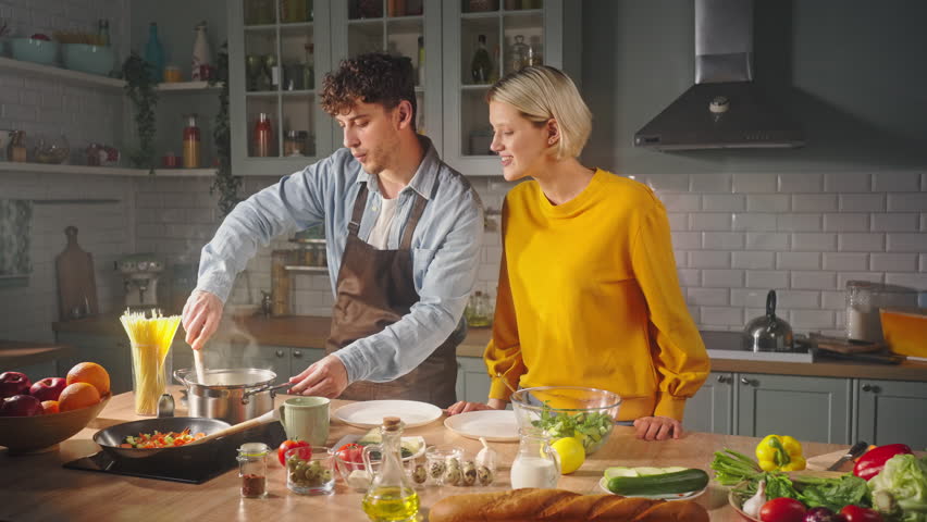 Charismatic bearded macho boiling Italian pasta, while cooking with his girlfriend in the home kitchen interior. Happy couple sharing happy moments together, enjoying preparing romantic dinner for two