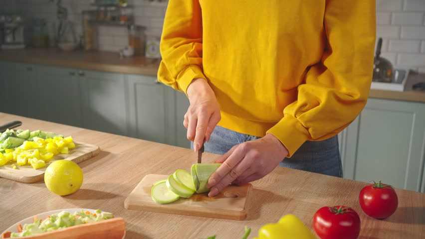 Selective focus on hands of a young woman, standing by wooden kitchen table and slicing vegetable marrow, preparing healthy vegan meal at home kitchen interior. Close-up of a couple cooking together Royalty-Free Stock Footage #1104213227