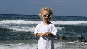 Small curly blonde Caucasian girl against background of sea waves washing shore. Fashionable girl stands in sun with her arms outstretched, smiling and enjoying rest holidays. Weekend on warm sea.