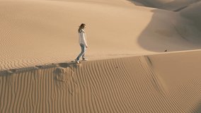 Slow motion 4K aerial drone footage of traveler woman leaving footprints on sand dune at golden sunset. Long shadows on golden sand California USA. Tourist female walking by desert natural landscape