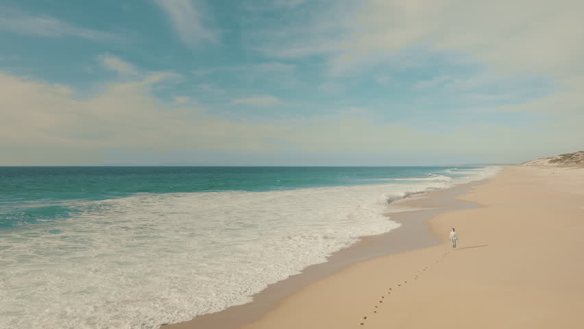 Aerial view of blonde woman walking barefoot by the blue ocean, leaving footprints in the sand. Foamy waves are rolling towards the sandy beach. Tropical summer vacation. 