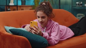 Preteen school girl texting share messages content on smartphone social media applications online, watching relax movie. Happy child kid uses mobile phone at home room apartment lying on orange couch