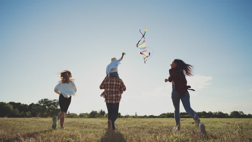 View from back of happy family running with kite flying in wind. Sports outdoors recreation in motion. Summer landscape. Parents play catch-up with children. Enjoy rest together. Leisure activity. Royalty-Free Stock Footage #1104230707