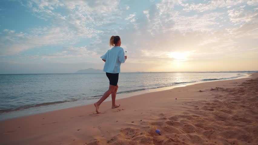 Barefooted woman jogging along sandy sea ocean beach holding bottle of water at sunset. Outdoors training, running, wellness sport training, workout of female. Active lifestyle, body care concept. Royalty-Free Stock Footage #1104230723