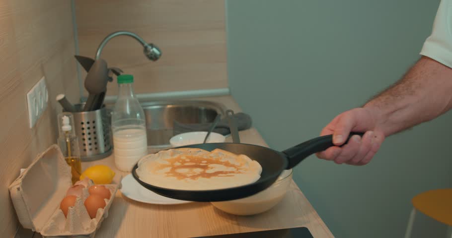 Man holds pan, flips a pancake in the kitchen. guy has fun with your food. Shot of a young man making pancakes at home. close up side view cropped video slow motion Royalty-Free Stock Footage #1104232999