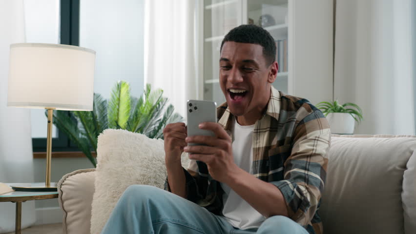 Happy excited African American guy user man winner celebrate online victory with mobile phone smartphone winning triumph win achieve on sofa at home cellphone prize bet achievement champion at couch Royalty-Free Stock Footage #1104235061
