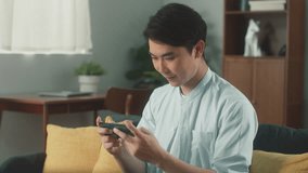 Happy Attractive Young Asian Man Hand Holding Smartphone Focus on Playing MOBA MMORPG Mobile Game Get MVP Victory Express Joyful Win Excited Face Feeling Cheerful Sit On Couch In Cozy Living Room