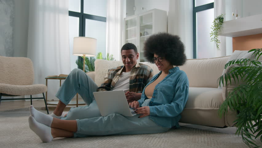 African American couple on floor at home using online laptop computer browsing shopping together pay bills choose products order delivery do high five gesture teamwork happy family in own apartment Royalty-Free Stock Footage #1104250399