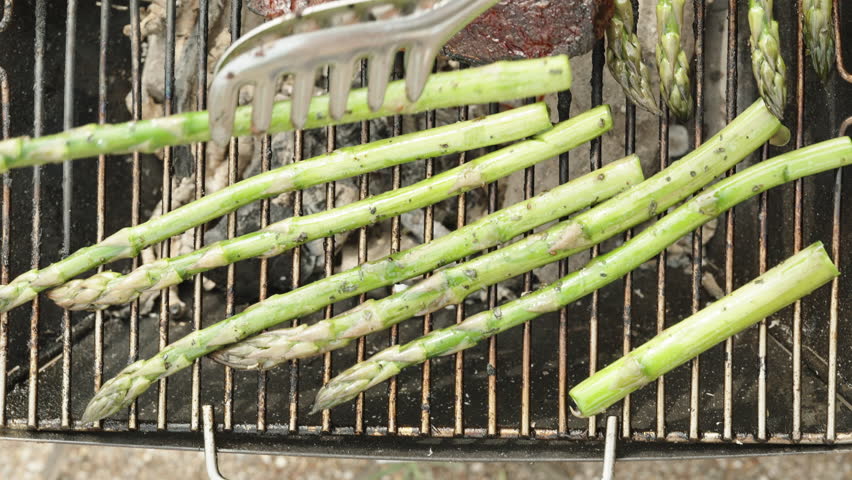 I place asparagus spears on the grill alongside the cooking steak. Top view. Slow motion.