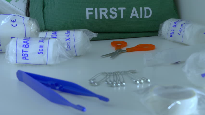 Closeup tilt shot of a green first aid kit bag, with various item in front ready to use – bandages, scissors, tweezers and safety pins.