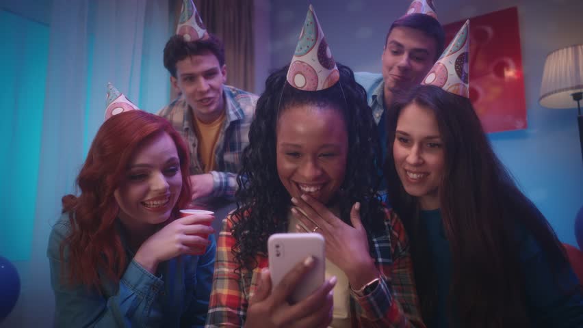 An African American girl shows the phone to her friends, they are emotionally looking at photos, videos or talking on a video call. A group of friends in party hats at a house party. Slow motion. Royalty-Free Stock Footage #1104256637