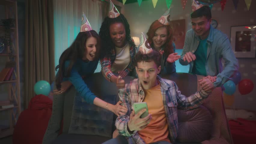 A guy in a party hat is playing an online game on his phone, his friends are watching him standing behind him. The guy wins, makes a victory gesture, accepts congratulations from friends. Slow motion. Royalty-Free Stock Footage #1104256699