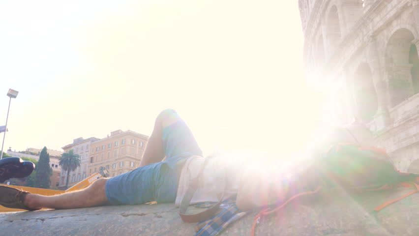 Happy young man tourist lying on a column with bike taking pictures with vintage camera at colosseum in Rome, Italy at sunrise.