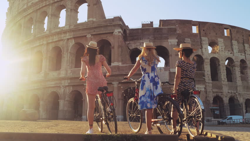 Three happy young women friends tourists with bikes waving hats at Colosseum in Rome, Italy at sunrise. Royalty-Free Stock Footage #1104256995