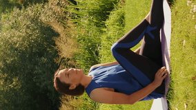 Young cute woman stretching on yoga mat near river in morning. Vertical video. Charming female in sportswear doing yoga exercises outdoors at sunset, enjoying nature and fresh air. Healthy lifestyle