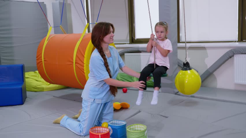 Child with physiotherapist on swing in sensory integration room. Physiotherapy and neuro corrections in children therapy center Royalty-Free Stock Footage #1104259611