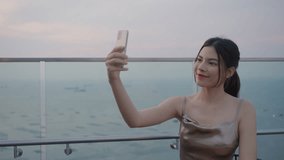 Beautiful woman in elegant golden dress in luxury bar on rooftop of city hotel near sea using smartphone cheerful selfie or take a photo or video call.