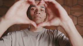 Young romantic kind African American man demonstrates figure of heart made of fingers to send video valentine to girlfriend or invite girl for date, stands indoors. Love, relationships