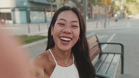 Pretty brunette girl, dressed in white top and jeans, smiles while taking selfie while sitting on bench on cityscape background, shows gesture of victory - Powered by Shutterstock - Get 15% off with code: PIKWIZARD15