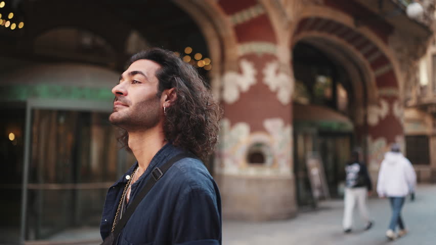 Young attractive italian guy with long curly hair and stubble looks into the camera and smiles at old buildings background. Stylish man with an earring in his ear and lot of chains