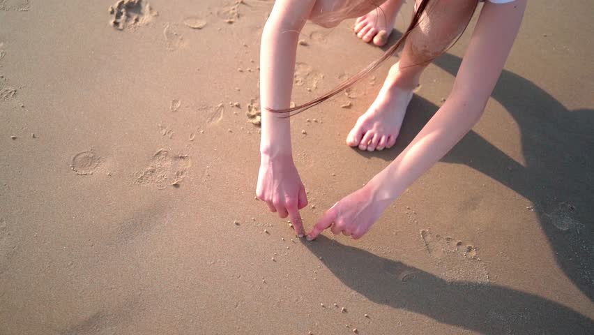 Girl hand draws symbol of heart and love on sandy beach. people play in sand on sunny summer day at sea coast. love of shore travel concept.  | Shutterstock HD Video #1104276035