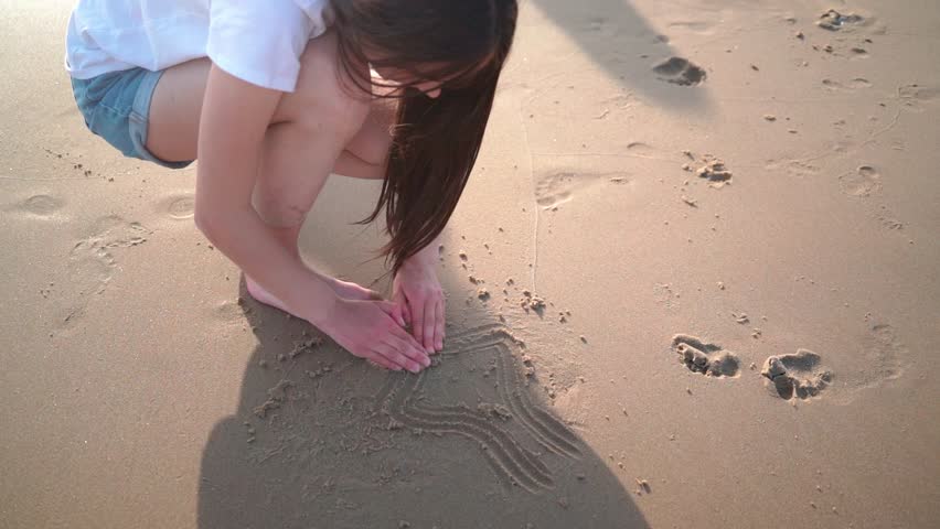 Girl hand draws symbol of heart and love on sandy beach. people play in sand on sunny summer day at sea coast. love of shore travel concept.  | Shutterstock HD Video #1104276037