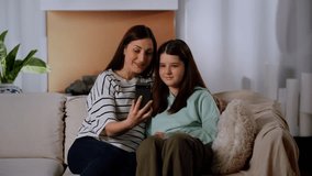Mom and daughter are sitting on the couch and talking with someone via video link through a smartphone. A little girl sits in the living room next to her mother and communicates with her father