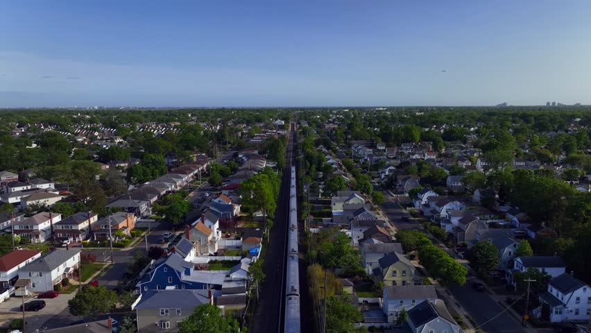 An aerial view of a Long Island railroad train travelling on a sunny day. The camera dolly in, centered over the tracks following the train running through residential neighborhood on Long Island, NY. Royalty-Free Stock Footage #1104279821