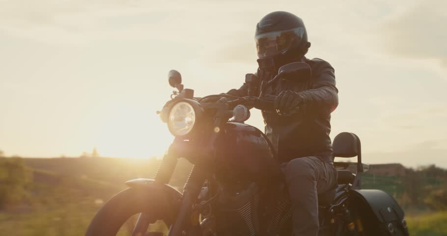 Man in a black leather jacket driving a black motorcycle through the dusk sky. Biker wearing a black helmet driving a vintage motorbike through a country highway. Optical flares of sun rays.