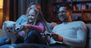 Portrait of parents with daughter playing video games at home. Happy family sitting on a sofa and playing on PlayStation using gamepads. Having fun concept