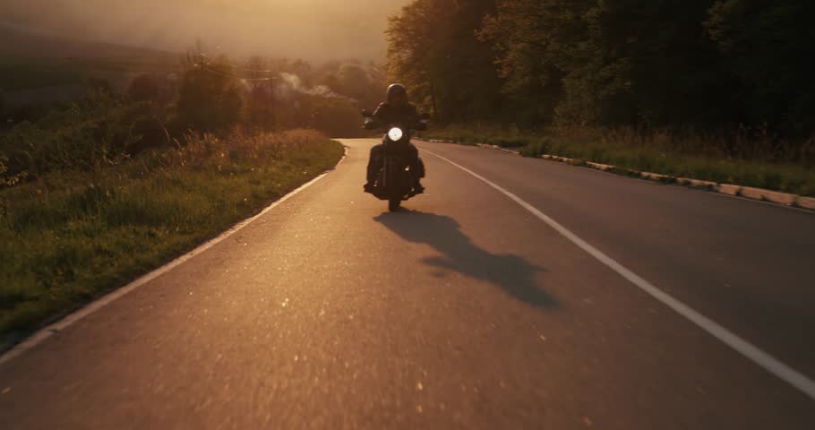 A man driving a motorbike through an empty highway during beautiful sundown. Biker guy driving a motorcycle with a head-light on through country road during sunset, wearing a leather jacket and helmet