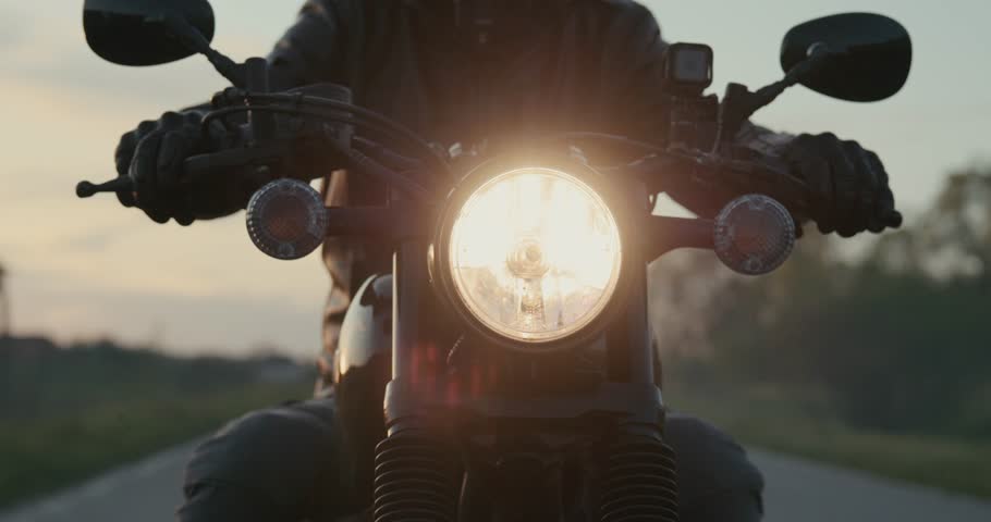 Portrait of a handsome man driving a classic motorcycle during a sunset. Biker wearing a black helmet and leather jacket riding a black vintage motorbike through country highway. Slow-motion shot