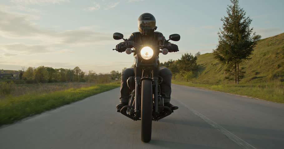 Slow-motion shot of a man riding a motorcycle through an empty road. Male biker in a black leather jacket and black helmet driving a classic motorbike through the country road during sunset Royalty-Free Stock Footage #1104288667