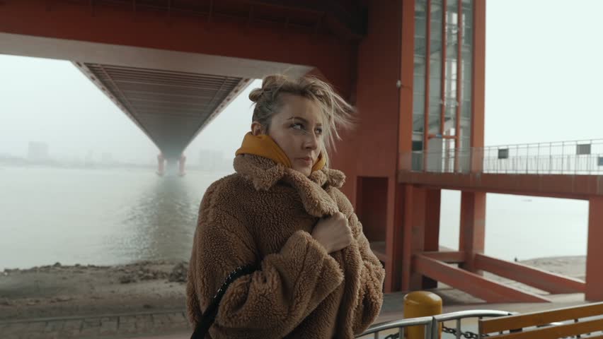 Blond Hair Girl in a Brown Coat Standing Under the Red Suspension Bridge, Cold Autumn Wind 