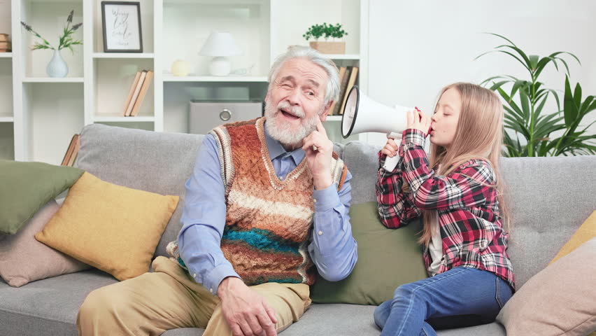 Grey-haired pensioner making funny face expressions while little restless girl shouting in white megaphone. Funky grandpa spending happy weekend with grandchild and enjoying spare time together. Royalty-Free Stock Footage #1104296677
